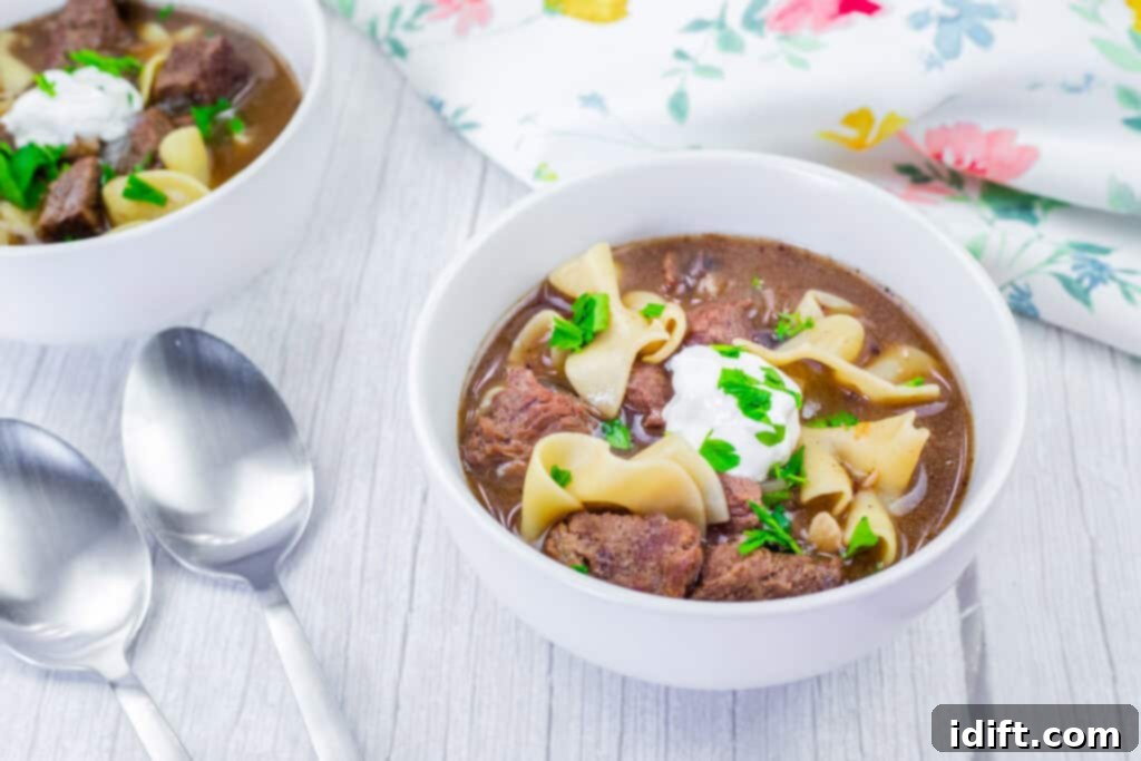Hearty Beef Stroganoff Soup 3 A warm bowl of Beef Stroganoff Soup, garnished with sour cream and parsley, sits on a rustic light wooden table alongside two spoons and a decorative floral napkin.