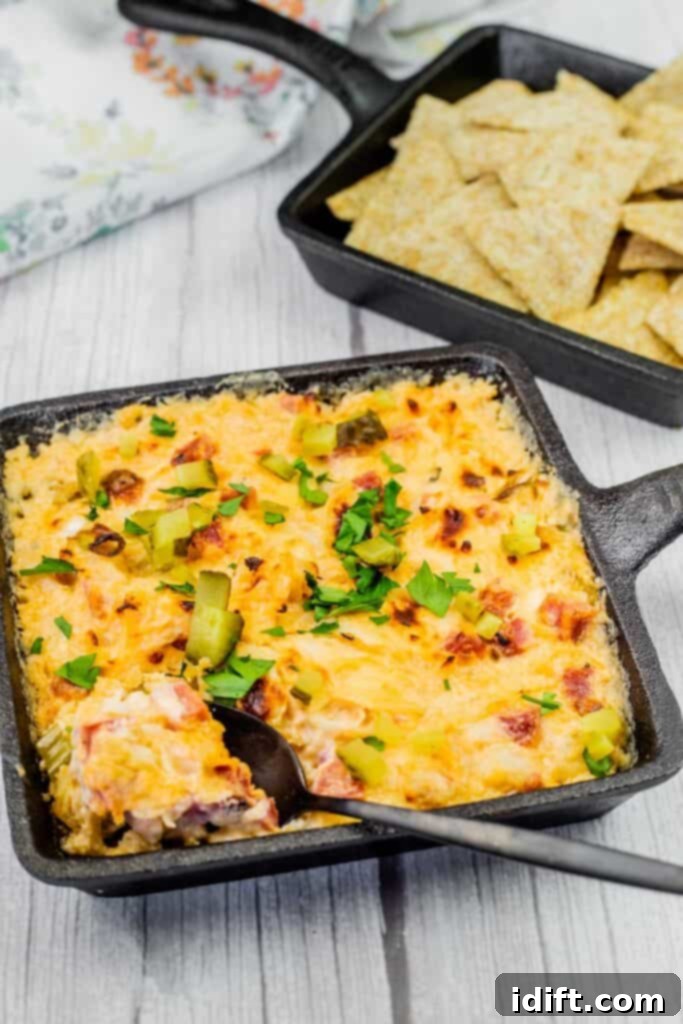 Golden Baked Reuben Dip 2 Hot Reuben dip in a cast-iron skillet, garnished with chopped pickles and fresh parsley, served with crispy crackers for dipping.