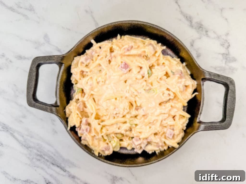 Golden Baked Reuben Dip 6 Top view of a creamy dish with diced ham and shredded cheese in a round black pan on a marble surface.