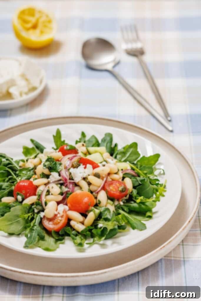 A White Bean Salad featuring arugula, cherry tomatoes, white beans, and red onions on a plate. A cut lemon, feta, and utensils are in the background on a checkered tablecloth.