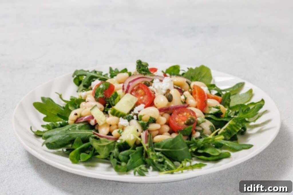 A White Bean Salad on a plate with arugula, cherry tomatoes, white beans, diced cucumber, red onion, and crumbled feta cheese.