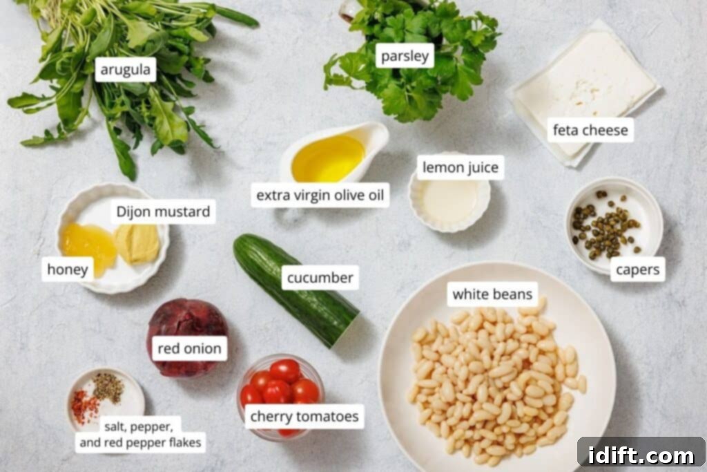 Ingredients for White Bean Salad arranged on a table: arugula, parsley, feta cheese, capers, lemon juice, extra virgin olive oil, Dijon mustard, honey, cucumber, white beans, red onion, cherry tomatoes, spices.