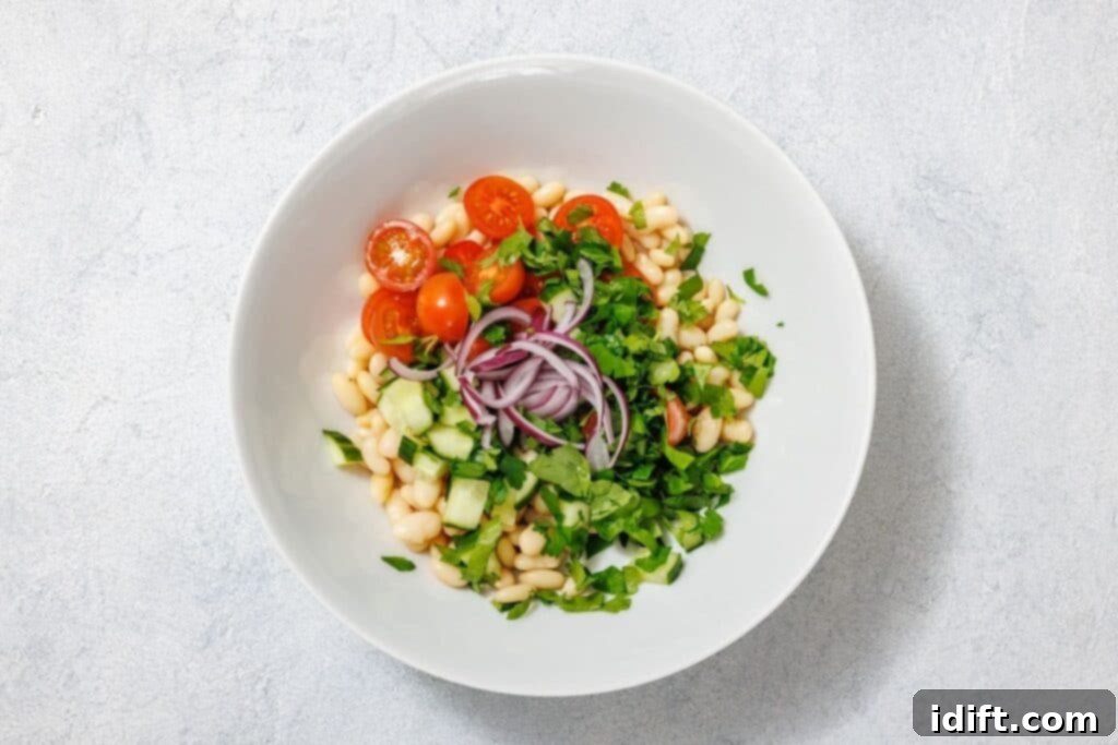 White bowl with a salad of white beans, cherry tomatoes, sliced red onions, cucumber, and chopped greens on a light gray background.