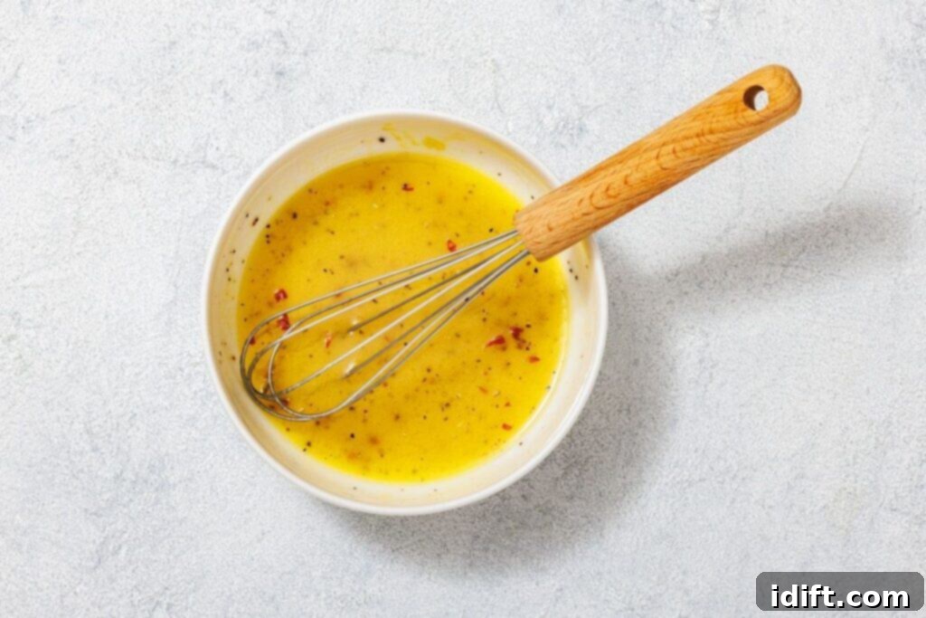 A bowl of yellow salad dressing with red flakes and a wooden-handled whisk on a light textured surface.