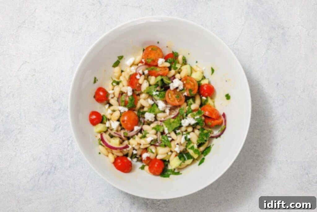 A bowl of salad with white beans, cherry tomatoes, cucumber, red onions, feta cheese, and herbs on a light gray background.