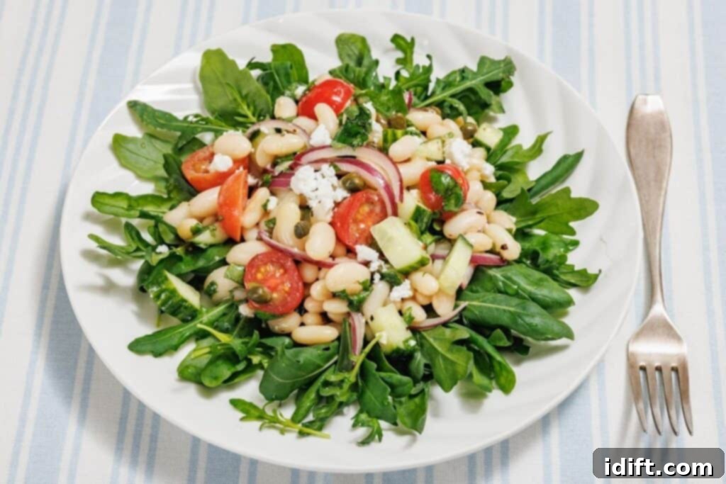 A white plate with a White Bean Salad featuring arugula, white beans, cherry tomatoes, red onions, cucumber, and crumbled cheese on a striped tablecloth, with a fork beside it.