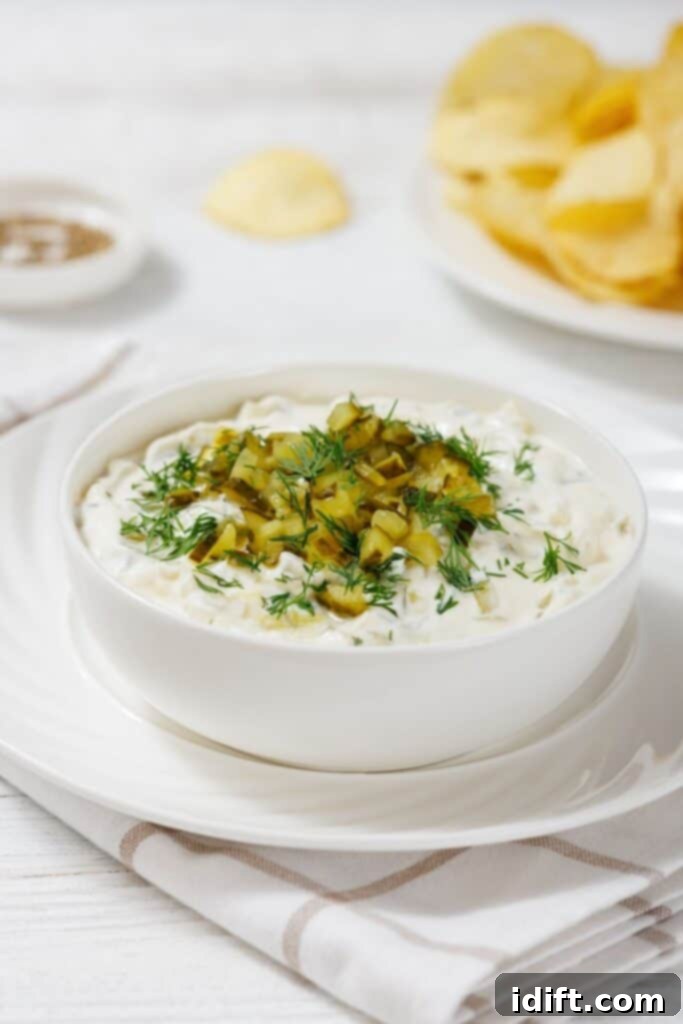A bowl of creamy Dill Pickle Dip topped with chopped pickles and dill, placed on a white saucer. A plate of potato chips is in the background on a white tablecloth.