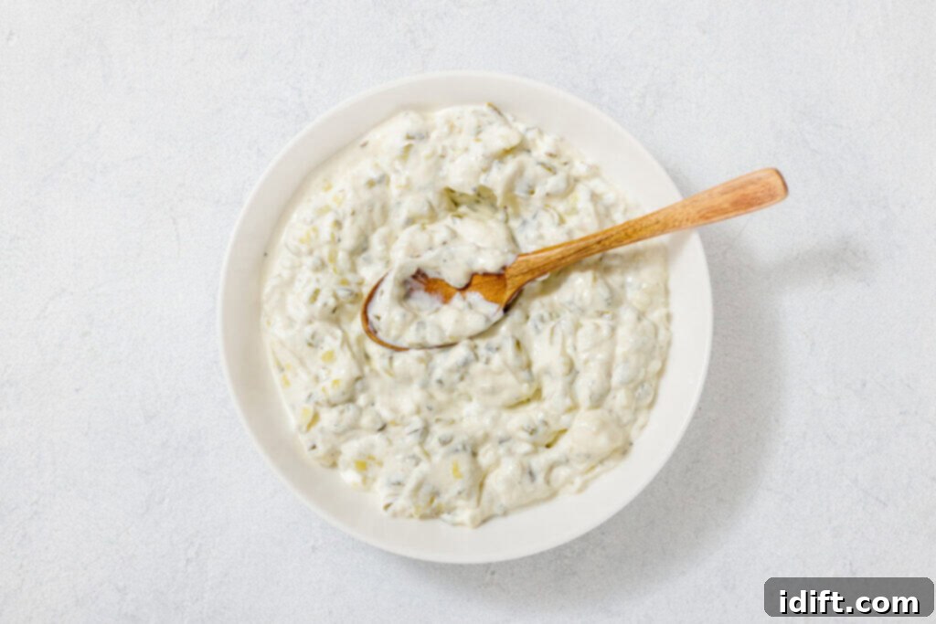 A bowl of creamy tzatziki sauce with herbs, shown from above. A wooden spoon rests in the sauce.