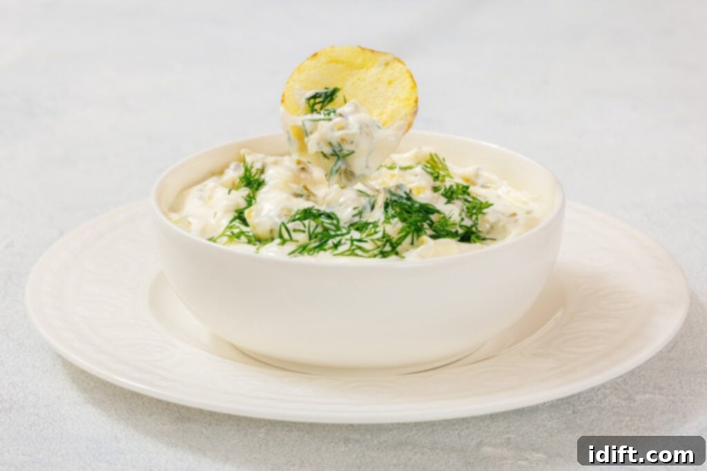 A bowl of creamy Dill Pickle Dip topped with chopped dill, with a potato chip dipped into it, placed on a white plate.