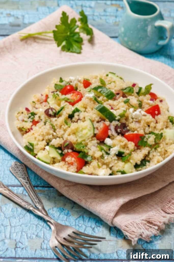 A beautifully arranged bowl of Mediterranean Quinoa Salad, featuring fluffy quinoa, bright red cherry tomatoes, sliced green cucumbers, dark Kalamata olives, and crumbled white feta cheese. The bowl rests on a rustic pink fabric, with a small jug and a sprig of fresh parsley in the softly blurred background on a light blue wooden surface. This visually appealing image highlights the freshness and colorful ingredients of the salad.