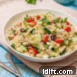 A bowl of quinoa salad with cucumbers, tomatoes, olives, and feta on a pink cloth. A small jug in the background and sprig of parsley on light blue wooden surface.