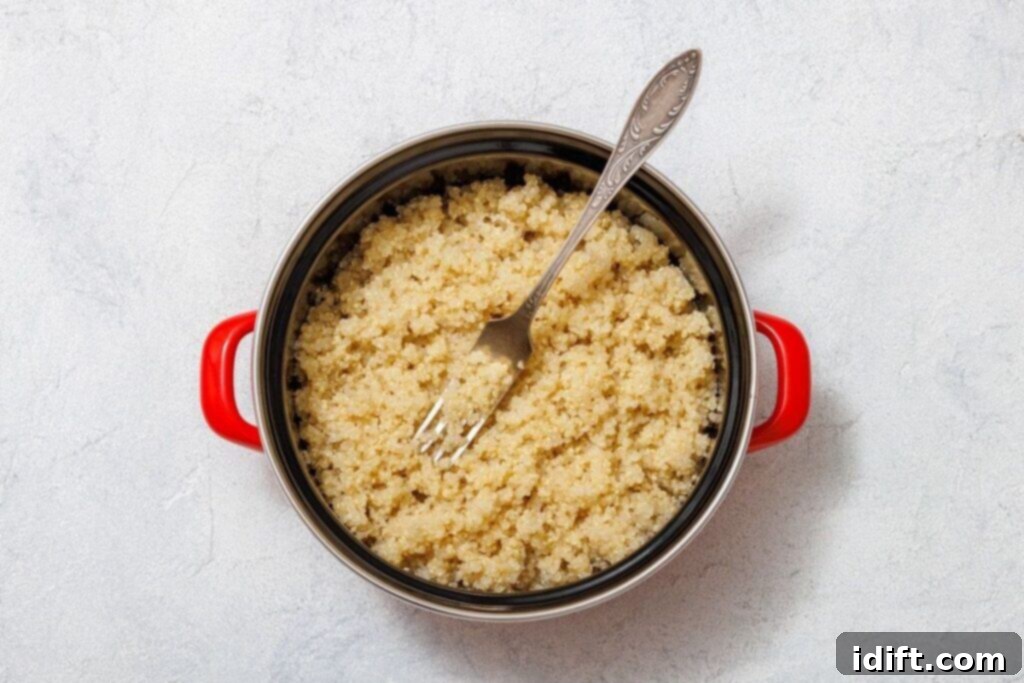 A red-handled pot filled with perfectly cooked, fluffy quinoa, with a metal fork resting inside, set on a light gray background. This image shows the quinoa after it has been cooked and fluffed.