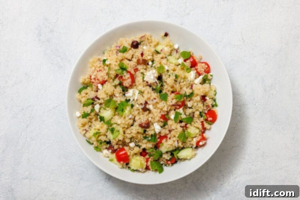 A large mixing bowl containing a colorful array of ingredients for the Mediterranean Quinoa Salad: cooked quinoa, bright cherry tomatoes, sliced cucumber, dark Kalamata olives, crumbled feta cheese, and fresh green herbs like mint and parsley. The bowl sits on a light textured surface, ready for the dressing.