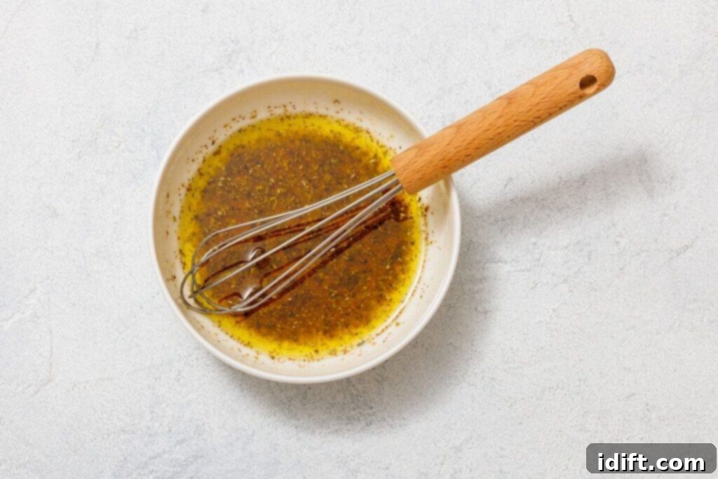 A small bowl holding a vibrant mixture of olive oil, lemon juice, red wine vinegar, dried oregano, and black pepper, with a small whisk resting inside. The bowl is placed on a clean white surface, highlighting the liquid ingredients of the dressing.