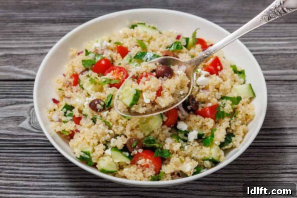 A large bowl of colorful Mediterranean Quinoa Salad, featuring cherry tomatoes, cucumbers, olives, feta, and fresh herbs, with a serving spoon resting inside. The salad is visibly dressed and ready to be tossed further or served. The bowl is placed on a light wooden surface.