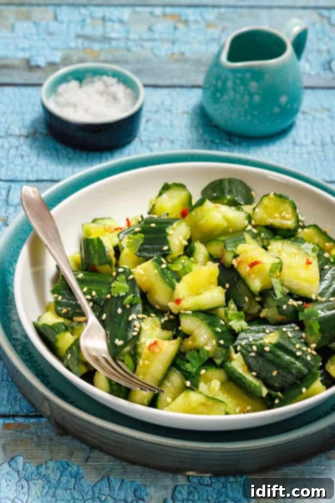 A beautifully presented bowl of Smashed Cucumber Salad, garnished with sesame seeds and fresh herbs, next to a fork on a rustic blue wooden table. A small salt dish and ceramic jug are visible in the soft background.