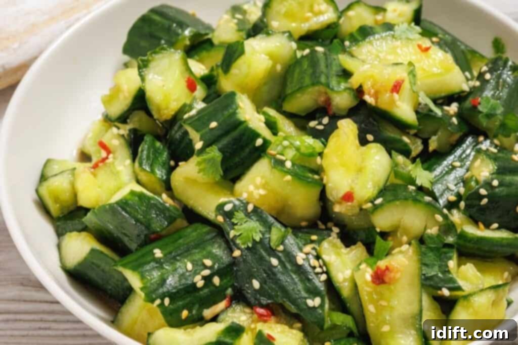 A beautifully presented serving of Smashed Cucumber Salad in a white bowl, generously garnished with toasted sesame seeds, vibrant fresh cilantro, and delicate red chili flakes, highlighting its appealing texture and color.