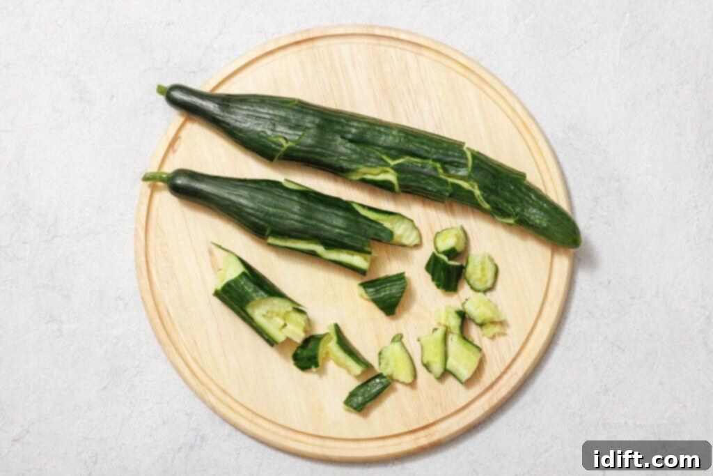Two English cucumbers, one partially peeled to show its texture, are placed on a round wooden board, with additional cucumber pieces already cut and scattered, demonstrating the initial preparation steps.