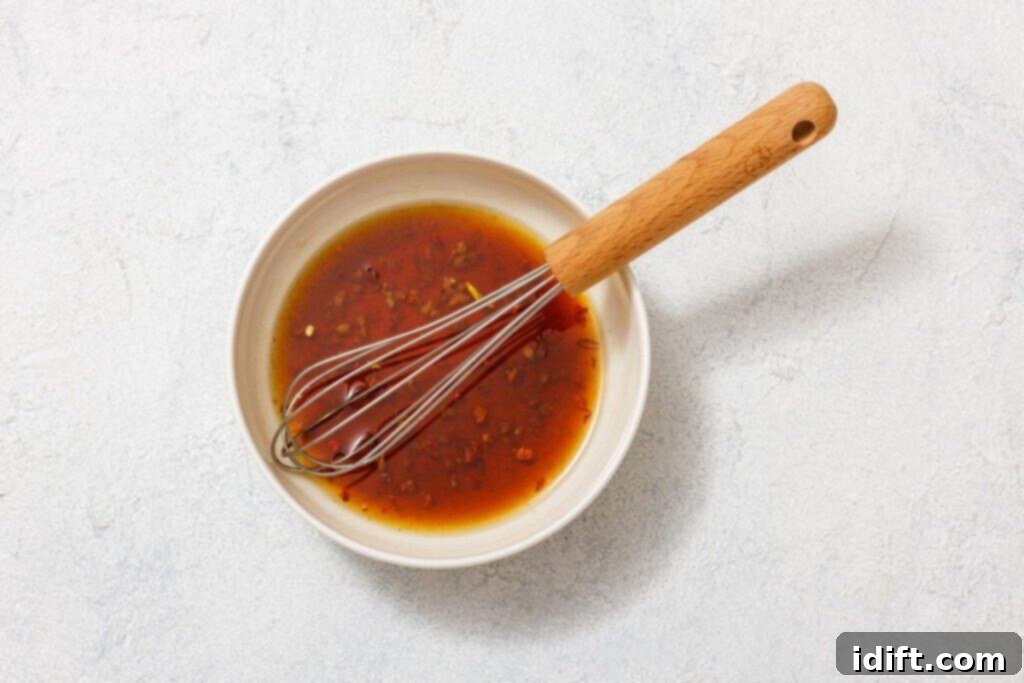 A small brown bowl containing a savory, rich dressing infused with finely chopped herbs, minced garlic, and vibrant red pepper flakes, accompanied by a whisk, resting on a textured light surface, showcasing the dressing preparation.