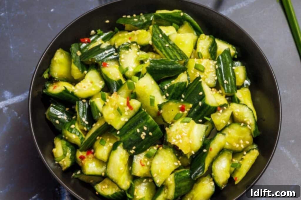 A vibrant bowl filled with smashed cucumbers, generously coated in the savory dressing and sprinkled with sesame seeds and fresh green herbs, illustrating the thorough mixing process on a dark countertop.
