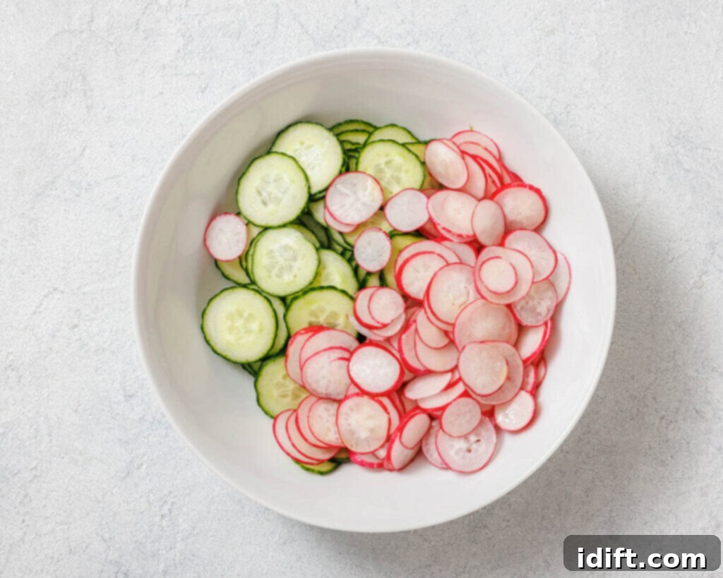 A white bowl with sliced cucumbers and radishes placed side by side on a light gray background.