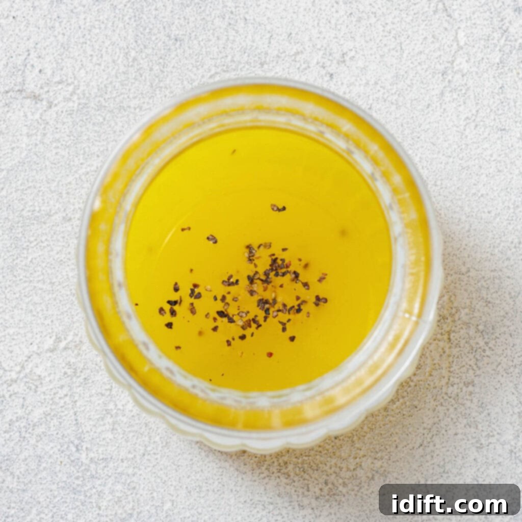 Top view of a transparent bowl filled with yellow liquid, possibly oil, topped with a sprinkling of black pepper on a light textured surface.