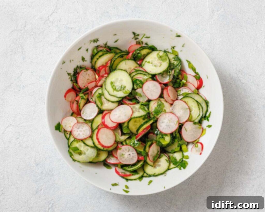 A bowl of fresh salad with sliced cucumbers, radishes, and herbs on a light surface.