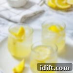 Perfectly Simple Lemonade 13 Three glasses of lemonade with ice and lemon slices on a white plate, with a bowl of sugar and sliced lemons in the background.