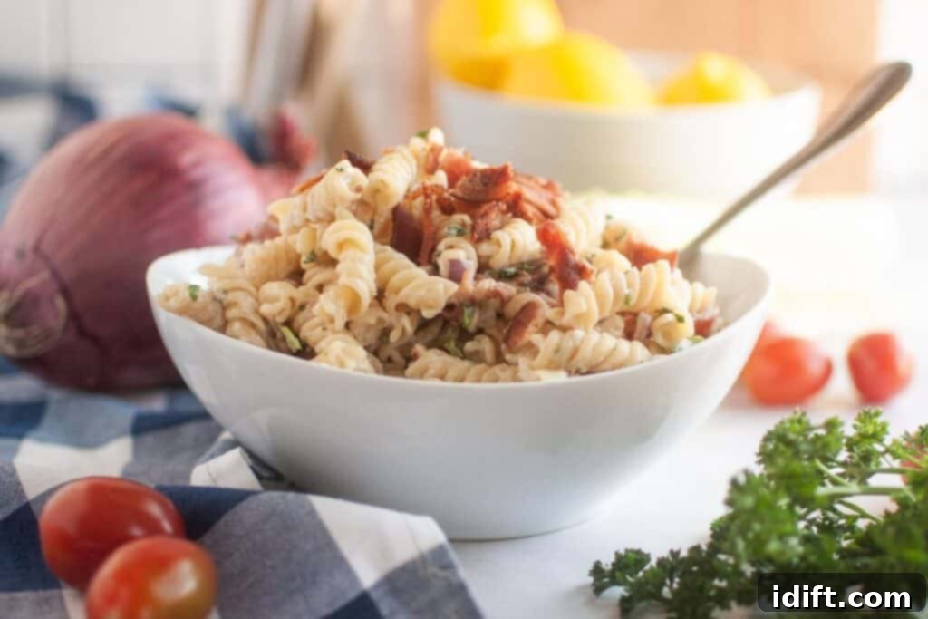 A white bowl filled with pasta salad, featuring spiral pasta, creamy dressing, pieces of bacon, and other visible ingredients, is shown with a spoon. Red onions, lettuce, cherry tomatoes, and parsley are in the background.