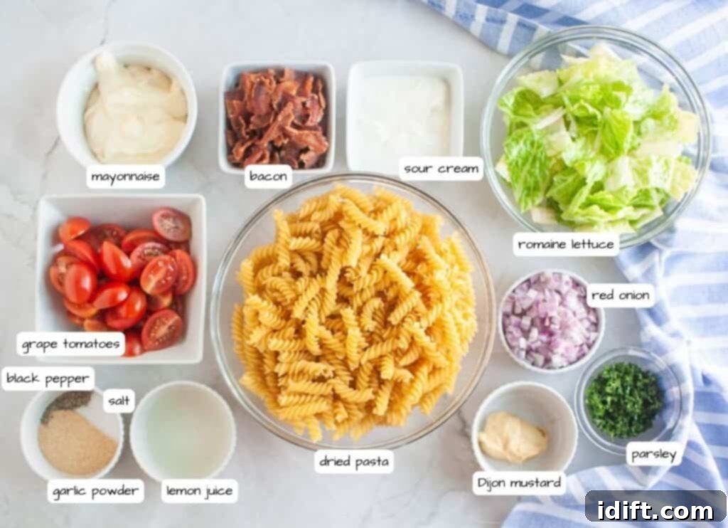 Overhead shot of various ingredients for a pasta salad, including cooked spiral pasta in a glass bowl, bowls of bacon, lettuce, red onion, parsley, tomatoes, and dressing components, arranged on a marble surface.