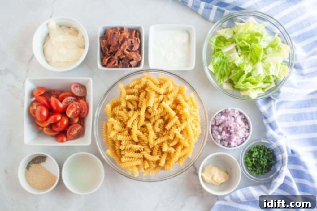 Overhead shot of various ingredients for a pasta salad, including cooked spiral pasta in a glass bowl, bowls of bacon, lettuce, red onion, parsley, tomatoes, and dressing components, arranged on a marble surface.