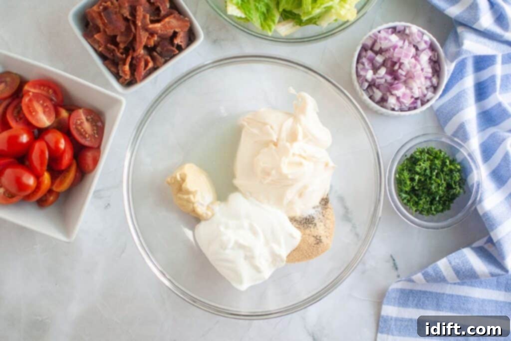 Overhead shot of a glass bowl containing creamy salad dressing ingredients, surrounded by bowls of bacon, lettuce, red onion, parsley, and tomatoes on a marble surface.