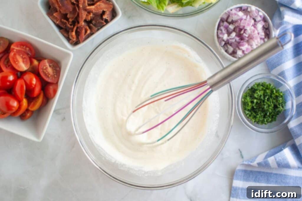 Overhead shot of a glass bowl containing mixed salad dressing with a whisk inside, surrounded by bowls of bacon, lettuce, red onion, parsley, and tomatoes on a marble surface.
