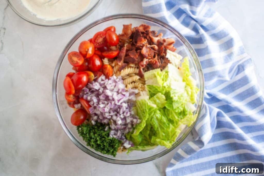 Overhead shot of a glass bowl containing BLT pasta salad ingredients, including pasta, tomatoes, bacon, lettuce, red onion, and parsley. A bowl of dressing and a blue striped cloth are also visible.