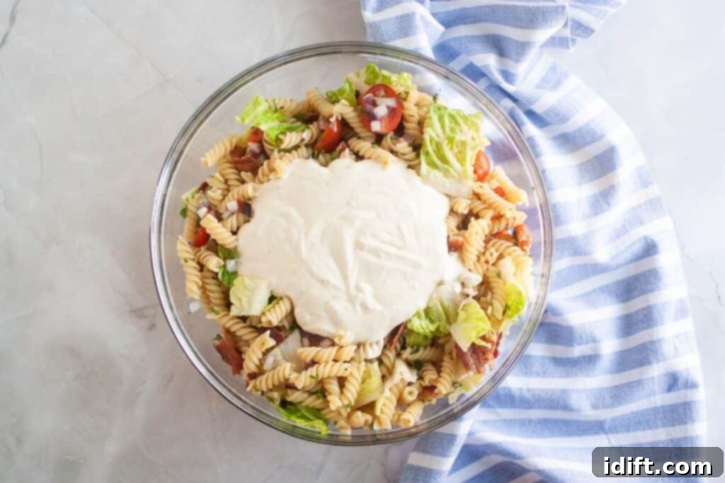 Overhead shot of a glass bowl filled with pasta salad, featuring spiral pasta, tomatoes, bacon, lettuce, and red onion, with a dollop of creamy dressing on top. A blue striped cloth is visible on the side.