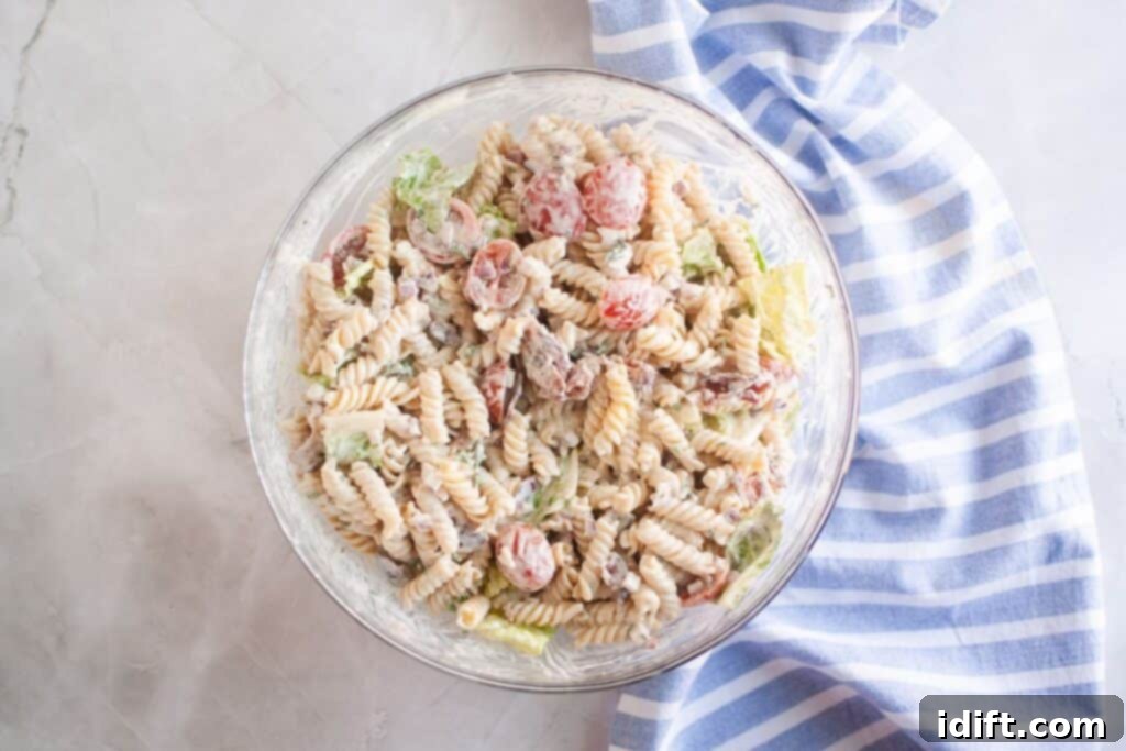 Overhead shot of a glass bowl filled with pasta salad, featuring spiral pasta, tomatoes, bacon, lettuce, and creamy dressing. A blue striped cloth is visible on the side.