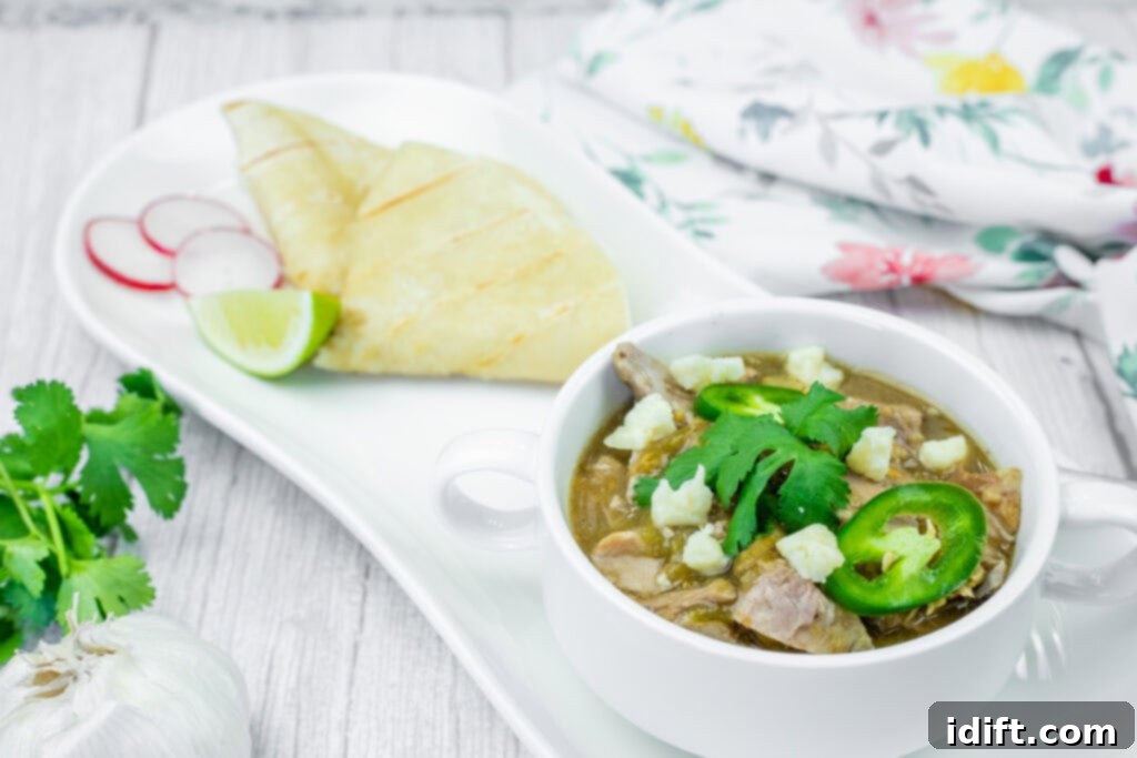 Tender Slow Cooker Pork Chile Verde 3 A white bowl of Slow Cooker Pork Chile Verde, featuring visible chunks of meat, white cheese crumbles, sliced jalapenos, and cilantro, sits on a white plate. Sliced radishes, a lime wedge, and folded tortillas are also on the plate. A head of garlic and cilantro sprigs are in the blurred background, along with a floral patterned cloth.