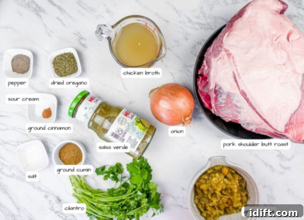 Tender Slow Cooker Pork Chile Verde 4 Overhead shot of various ingredients laid out on a white marble surface with gray veins, including a large piece of raw pork on a black plate, a jar of green salsa, a yellow onion, a measuring cup of broth, small bowls of spices, and fresh cilantro. Each ingredient is labeled with text.