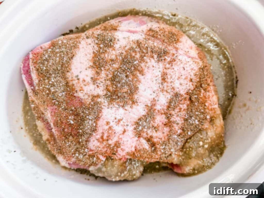 Tender Slow Cooker Pork Chile Verde 7 Overhead shot of a raw pork roast seasoned with spices, sitting in a white slow cooker with a thin liquid at the bottom. The pork has a pinkish hue and is coated with a visible blend of spices.
