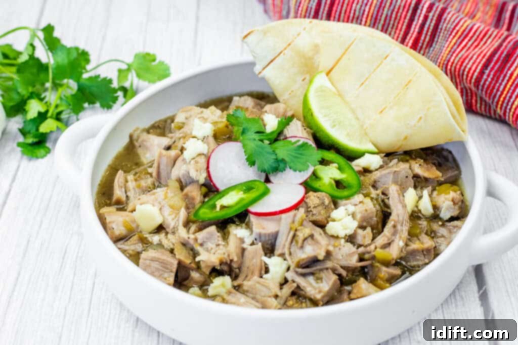 Tender Slow Cooker Pork Chile Verde 8 A white bowl of Slow Cooker Pork Chile Verde, featuring visible chunks of meat, radish slices, jalapeno slices, cilantro, and cheese, sits on a white wooden table. A folded tortilla and lime wedge are placed beside the bowl. A head of garlic and cilantro sprigs are in the background, along with a red and white striped cloth.