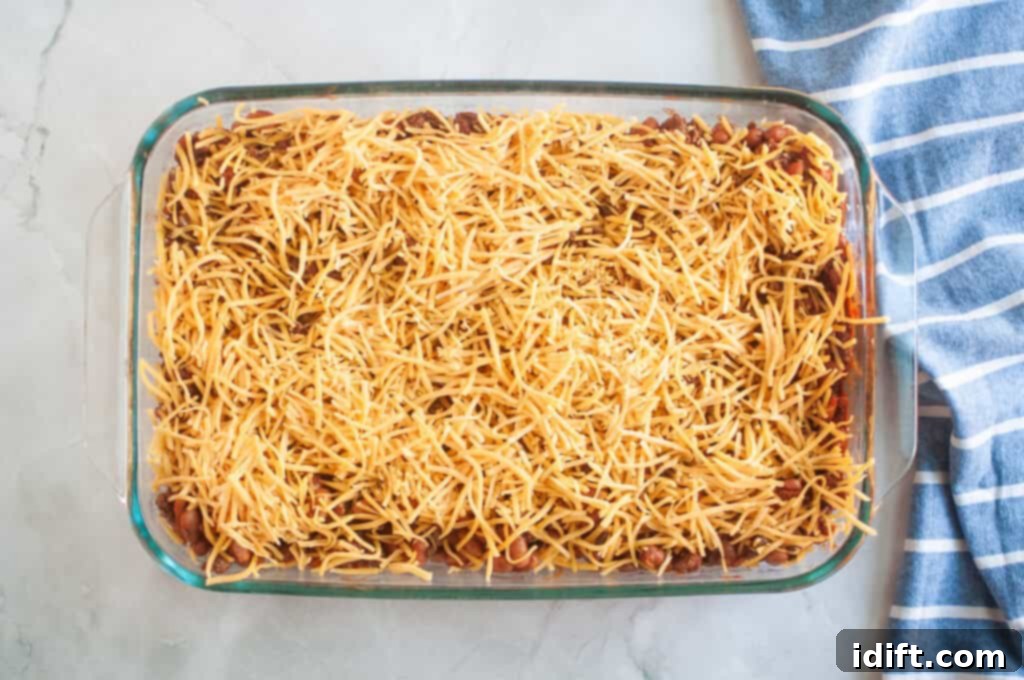 Overhead shot of a casserole with beans, ground beef, and shredded cheese in a clear glass baking dish, with a blue striped cloth beside it.
