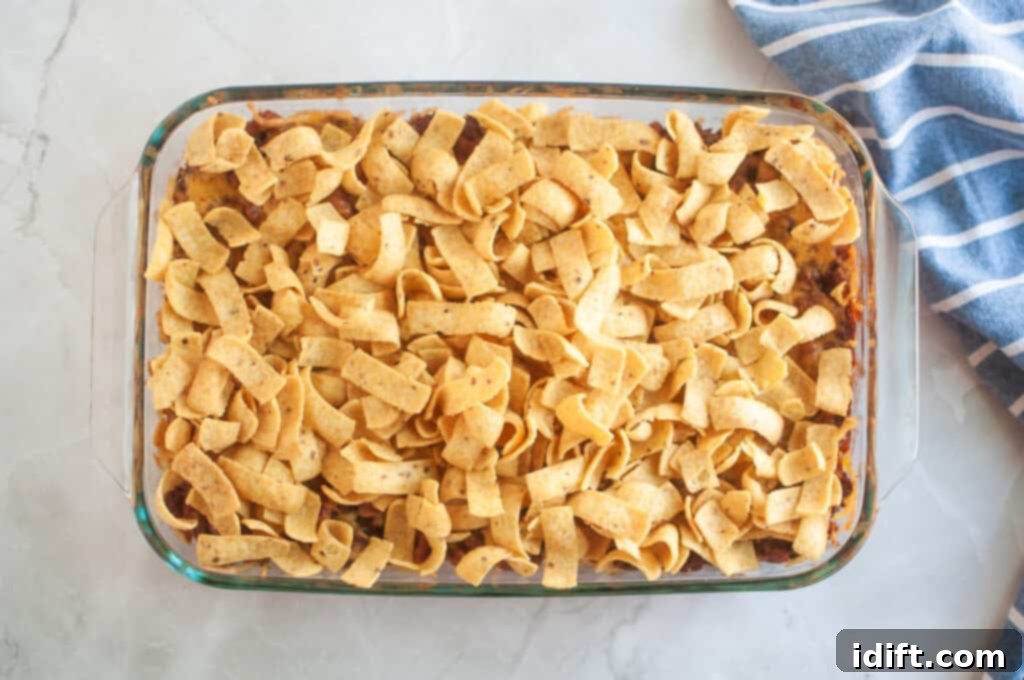 Overhead shot of a casserole with beans, ground beef, cheese, and chips in a clear glass baking dish, with a blue striped cloth beside it.
