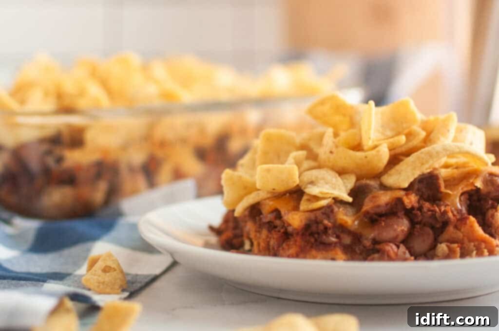A white plate with a serving of Frito pie, topped with corn chips, is shown on a light surface.