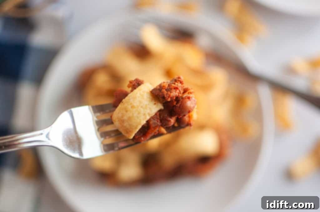 A close up take of Frito Pie in a fork with a blurry take of a small serve in a white plate in the background.
