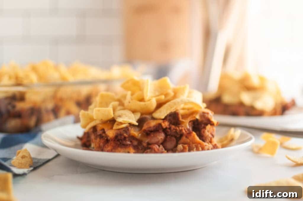 A front take of Frito Pie in a small white plate on a white table with corn chips on top and scattered.