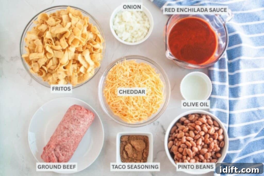 Overhead shot of ingredients for Frito pie, including Fritos, onions, enchilada sauce, cheese, ground beef, spices, oil, and beans, arranged on a light surface with a blue striped cloth and labels for each ingredient.