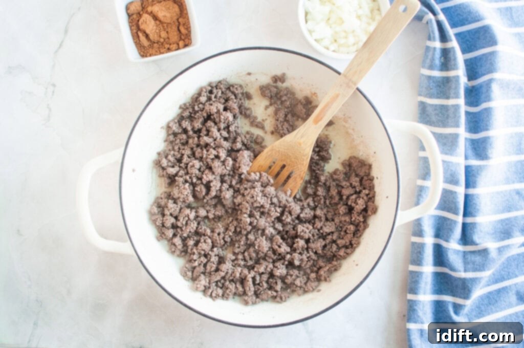 Overhead shot of cooked ground beef in a white pot with a wooden spoon, with bowls of spices and onions nearby.
