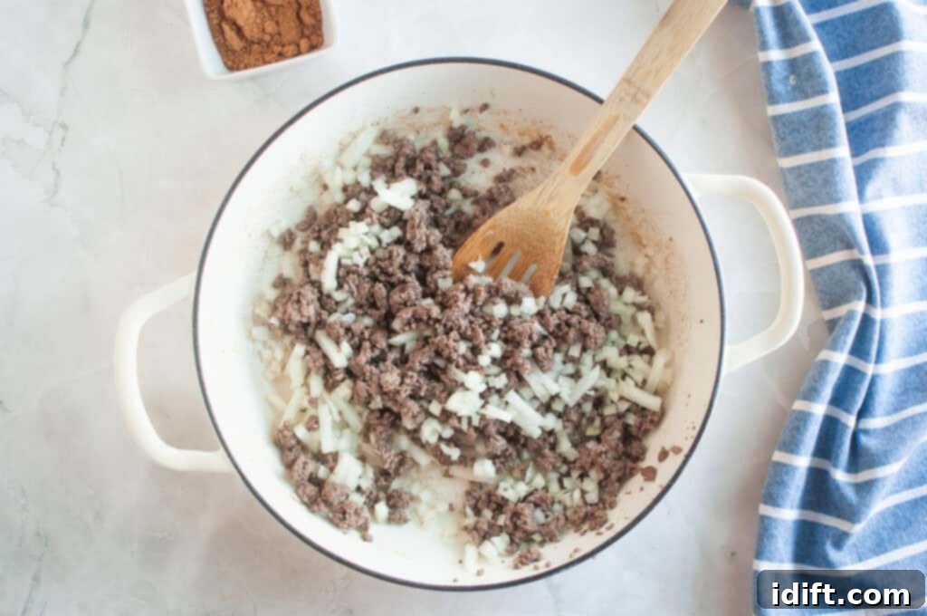 Overhead shot of cooked ground beef and chopped onions in a white pot with a wooden spoon, with a bowl of spices nearby.