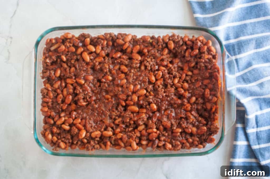 Overhead shot of baked beans and ground beef mixture in a clear glass baking dish, with a blue striped cloth beside it.