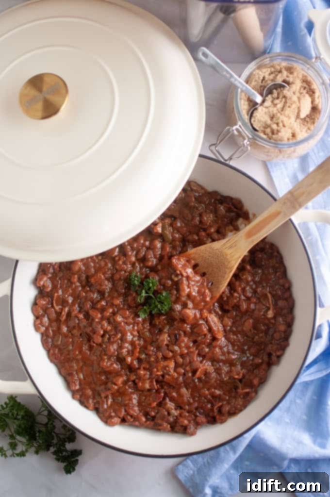 A white Dutch oven with baked beans topped with parsley sits on a marble surface next to a jar of brown sugar, a wooden spoon, and a blue cloth. This rich, smoky baked beans casserole is ready to be served.